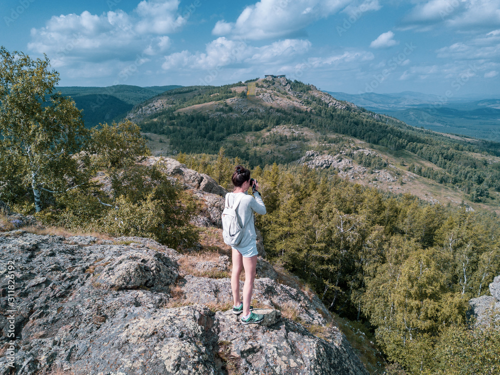 Young brunette girl stands on mountain top , takes photo of beautiful Ural landscapes; rocky cliff in summer sunny day