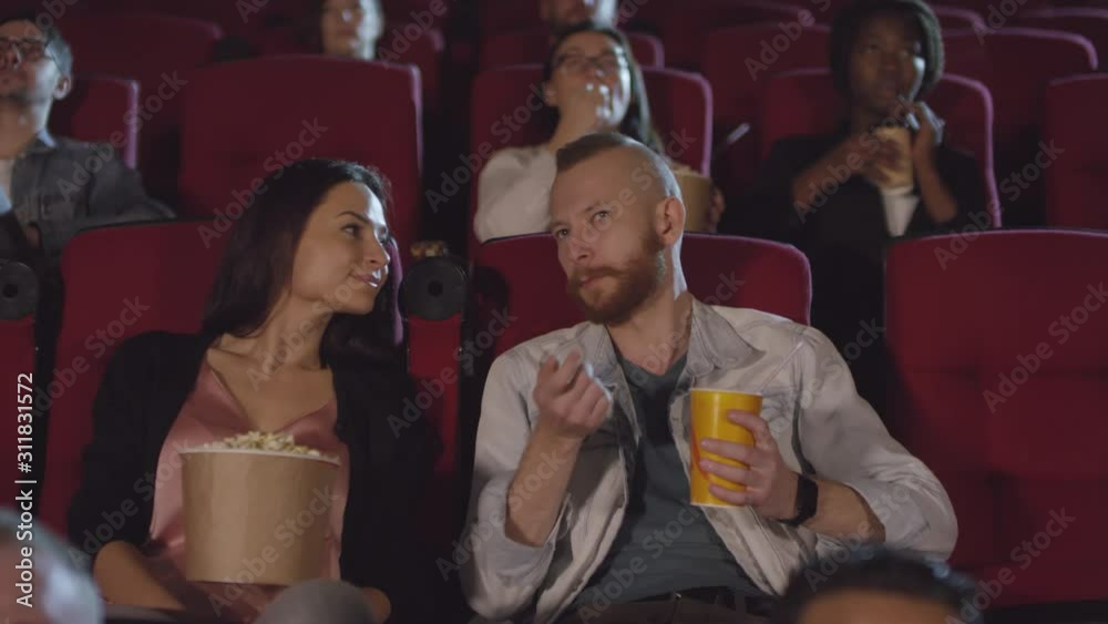 Tracking shot of cheerful couple with popcorn and soda drink watching movie in cinema and discussing it Tracking shot of cheerful couple with popcorn and soda drink watching movie in cinema and discussing it