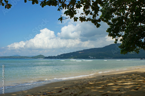 Fototapeta Naklejka Na Ścianę i Meble -  Coast of the Andaman sea. Sandy beach in the shade of a tree. Light waves roll ashore. Mountains in the background. A feeling of warmth and calm. Landscape from under a tree branch