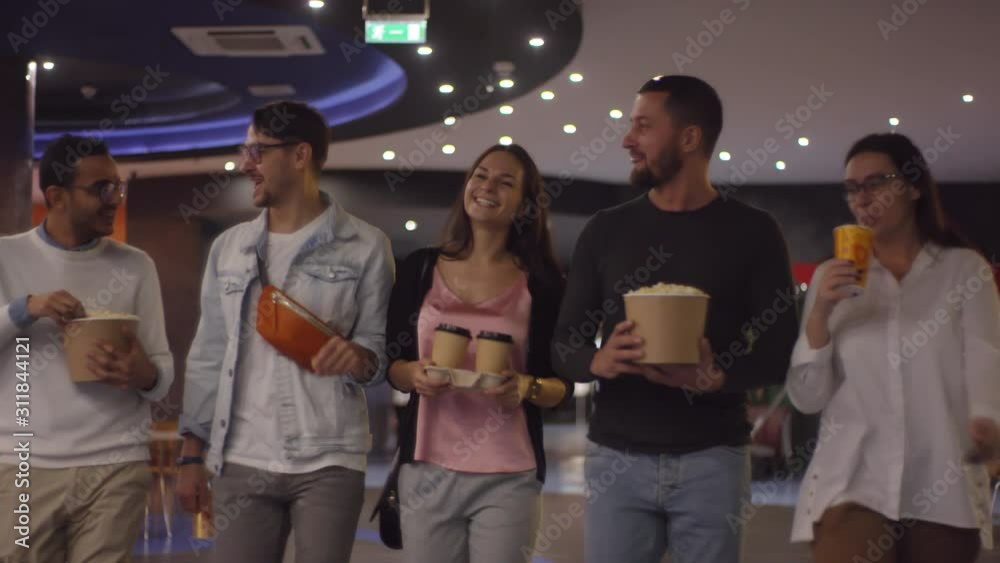 Tracking shot of happy friends eating popcorn and chatting while walking to watch movie at cinema