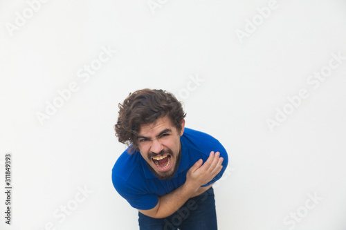 Frustrated unhappy guy screaming with pain, holding stomach, bending. Handsome bearded young man in blue casual t-shirt posing isolated over white background. Bellyache concept
