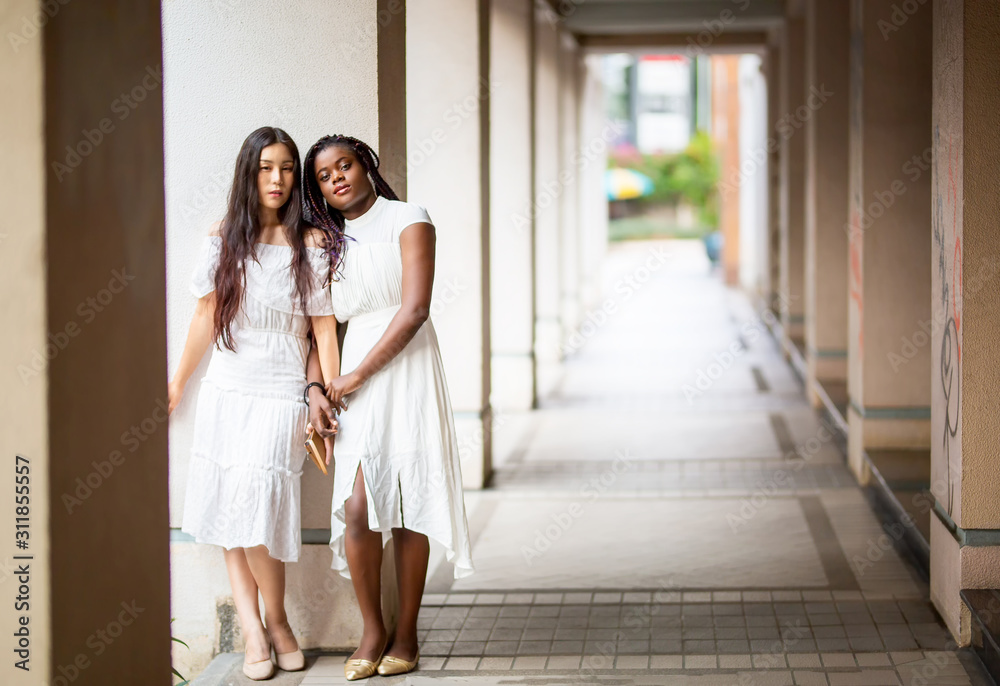 Fototapeta premium portrait of Asian and African american women in white dress against street