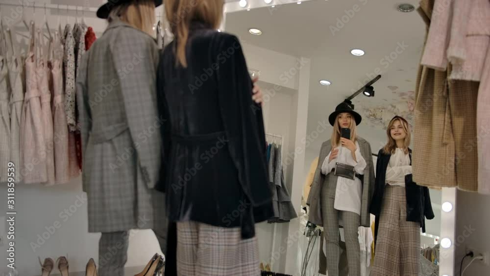 Pan left from below back view of happy millennial ladies in stylish elegant clothes taking selfie and looking at self in large mirror, trying on new clothes in clothing store in slow motion. Pan left from below back view of happy millennial ladies in stylish elegant clothes taking selfie and looking at self in large mirror, trying on new clothes in clothing store in slow motion.