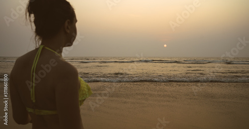 Anonymous woman near sea during sunset. Back view of unrecognizable female in swimwear standing on beach near waving sea in evening on resort