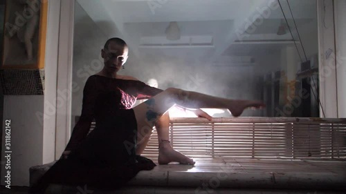 A mysterious young woman with shaved head sits on a sofa, swinging her dress and legs over to the side, with a harch light behind her. photo