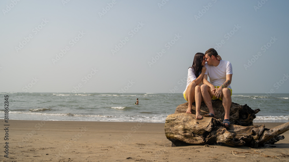 Happy couple sitting on driftwood near sea. Loving couple hugging and touching noses while sitting on driftwood during date on beach against waving sea and cloudless sky
