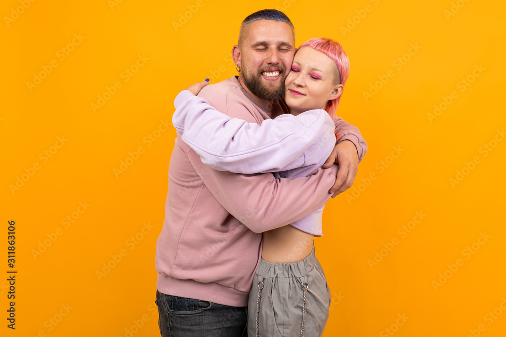 Unusual pretty woman with short pink hair and tattoo hugs with her boyfriend isolated on orange background Unusual pretty woman with short pink hair and tattoo hugs with her boyfriend isolated on orange background