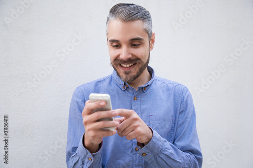Cheerful man with smartphone rejoicing at message, reading funny content, smiling. Grey haired young man in blue casual shirt posing isolated over white background. Happy cellphone user concept