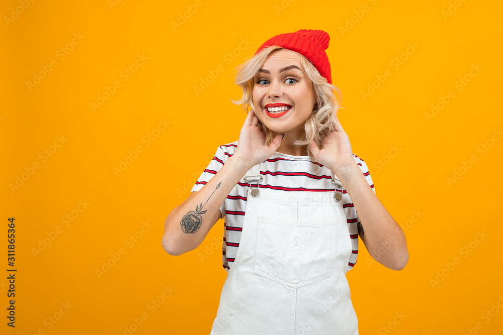 Beautiful young woman with short blonde curly hair and bright makeup in white overalls and red hat gesticulated and smiles, portrait isolated on orange background Beautiful young woman with short blonde curly hair and bright makeup in white overalls and red hat gesticulated and smiles, portrait isolated on orange background