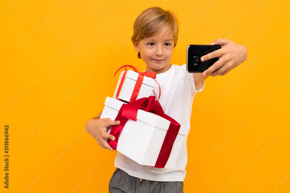 Caucasian boy holds many white boxes with gifts and do selfie, portrait isolated on yellow background Caucasian boy holds many white boxes with gifts and do selfie, portrait isolated on yellow background