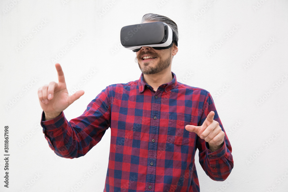 Content man using virtual reality headset. Cheerful bearded man in checkered shirt moving hands and using VR headset on grey background. Technology concept Content man using virtual reality headset. Cheerful bearded man in checkered shirt moving hands and using VR headset on grey background. Technology concept