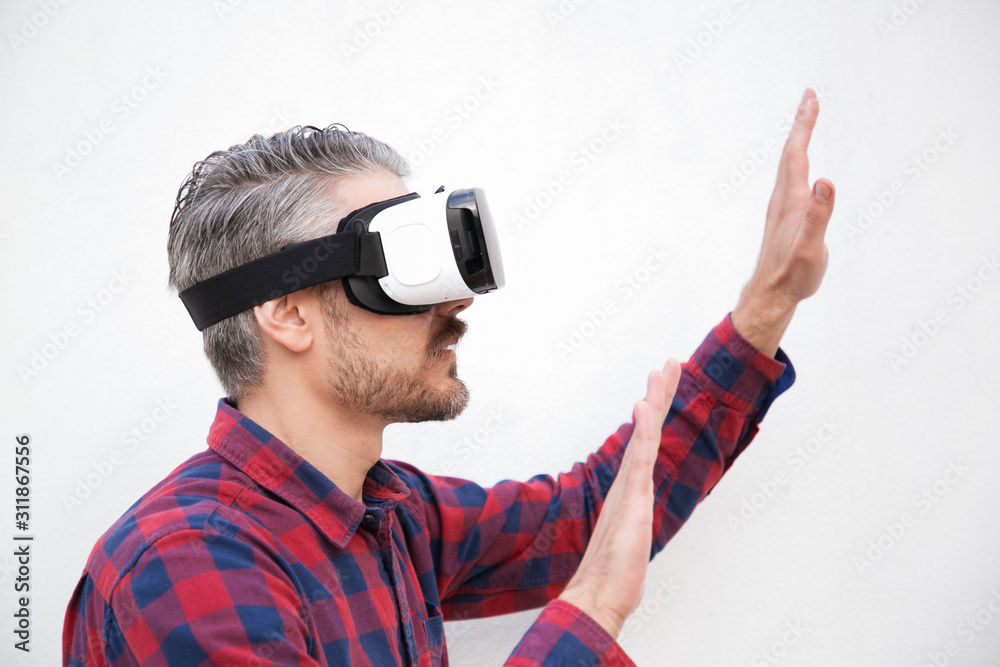 Focused man in VR headset moving hands. Side view of serious bearded man using virtual reality headset on grey background. Technology concept Focused man in VR headset moving hands. Side view of serious bearded man using virtual reality headset on grey background. Technology concept