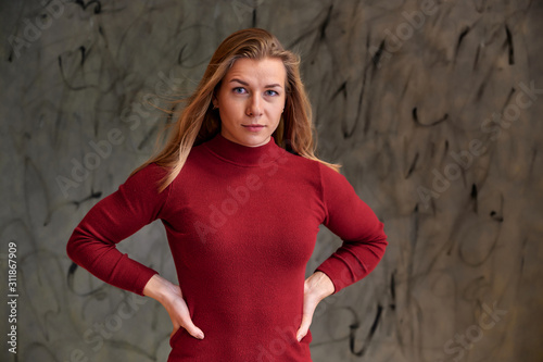 Portrait of a pretty fitness blonde girl with a beautiful smile in a burgundy dress on a gray alternative background. Cute looks at the camera, standing straight.