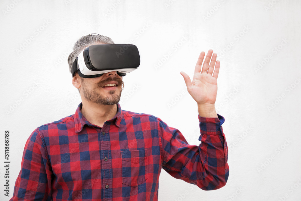Smiling man in VR headset moving hand. Handsome happy bearded man in checkered shirt moving with hand and using virtual reality headset on grey background. Technology concept Smiling man in VR headset moving hand. Handsome happy bearded man in checkered shirt moving with hand and using virtual reality headset on grey background. Technology concept