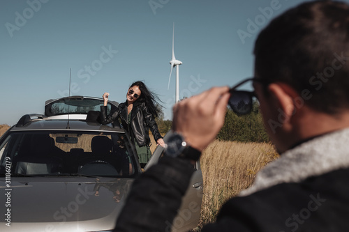 Happy Traveling Couple Enjoying a Car Trip on the Field Road with Electric Wind Turbine Power Generator on the Background
