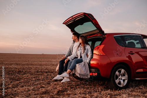 Young Happy Couple Dressed Alike in White Shirt and Jeans Sitting at Their New Car Trunk, Beautiful Sunset on the Field, Vacation and Travel Concept