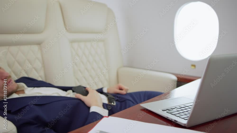 Exhausted middle aged businessman in suit sleeping on comfortable seat of private business jet with cell phone in his hands, open laptop and documents on desk