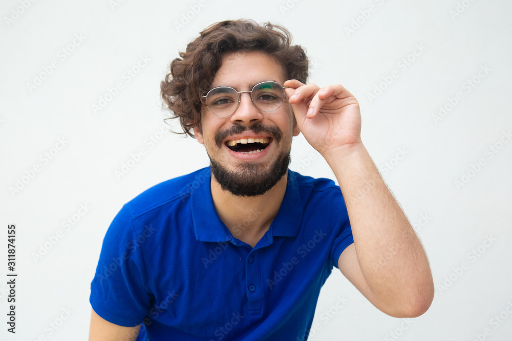 Happy cheerful satisfied guy touching glasses and staring at camera. Handsome bearded young man in blue casual t-shirt posing isolated over white background. Good news concept