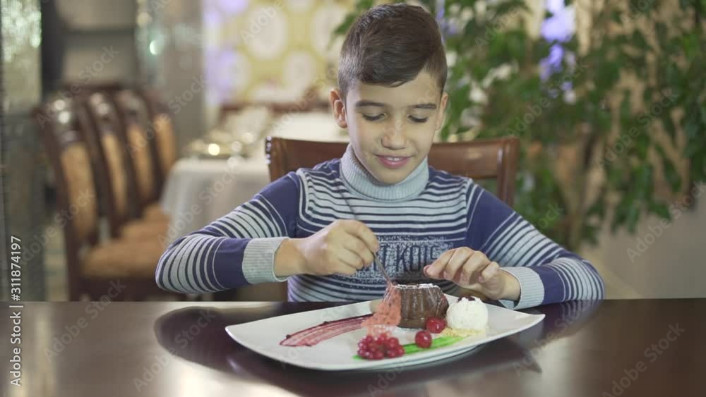 Cute little brunette boy enjoy eating chocolate dessert messy hands mouth in fancy restaurant cozy atmosphere background