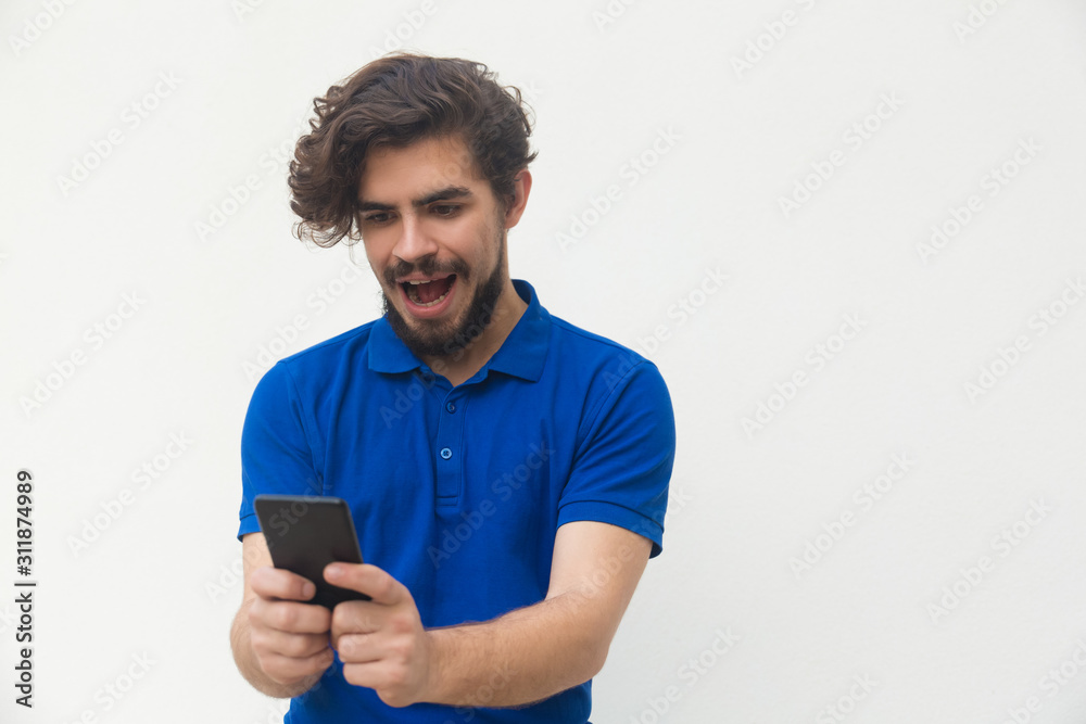 Happy surprised guy using smartphone, reading message shouting for joy. Handsome bearded young man in blue casual t-shirt posing isolated over white background. Good news concept