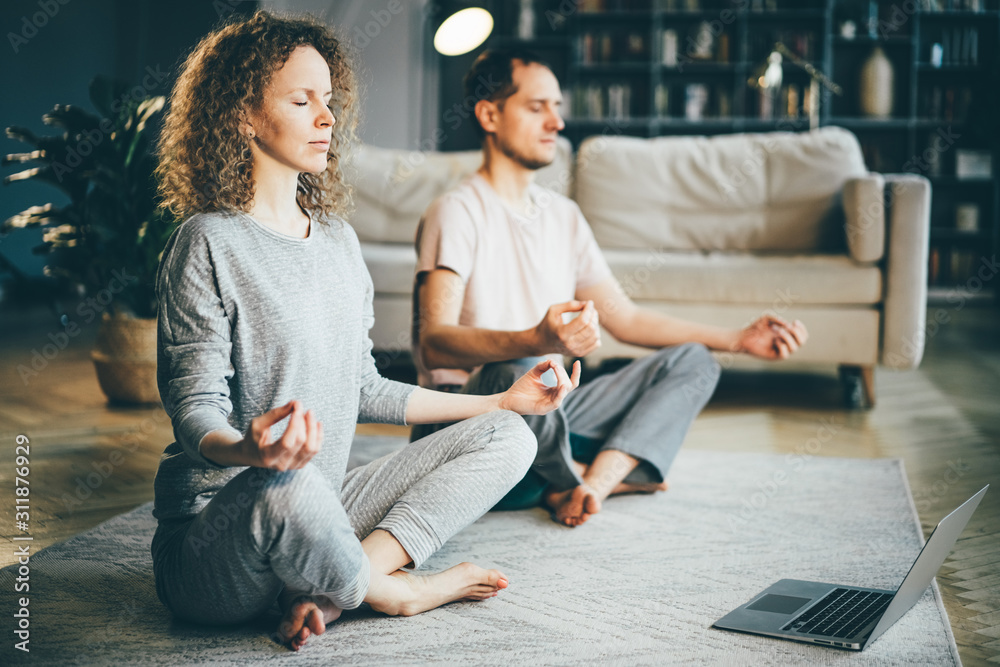 Calm couple in pajamas meditating, listening spiritual practices lessons on laptop, sitting on lotus pose at home. Yoga concept. - obrazy, fototapety, plakaty 