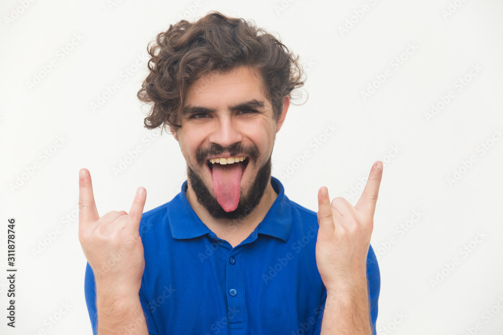 Crazy cheerful rocker sticking out tongue, making devil horns. Handsome  bearded young man in blue casual t-shirt posing isolated over white  background. Rock-n-roll fan concept Photos | Adobe Stock