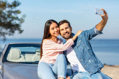 young couple taking selfie while sitting on the convertible together