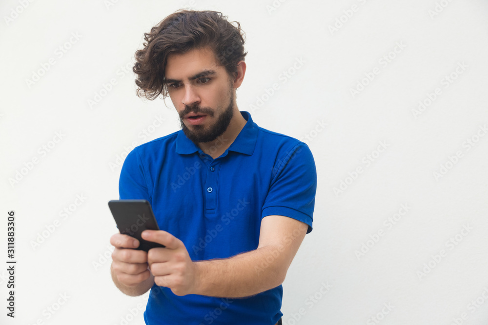 Shocked guy with smartphone reading message or watching content. Handsome bearded young man in blue casual t-shirt posing isolated over white background. Mobile phone using concept Shocked guy with smartphone reading message or watching content. Handsome bearded young man in blue casual t-shirt posing isolated over white background. Mobile phone using concept