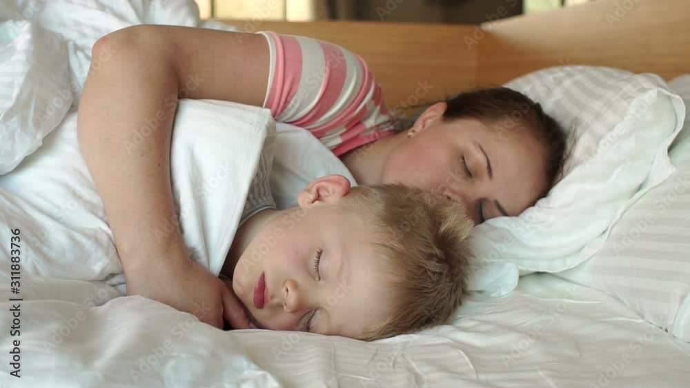 Close-up of mom gently hugging her sleeping son in the morning in bed. Mother and child sleep together in a white bed. Slow motion. Close-up of mom gently hugging her sleeping son in the morning in bed. Mother and child sleep together in a white bed. Slow motion.