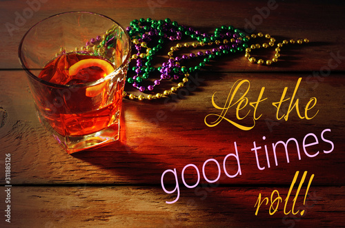 Close-up view of sazerac cocktail with a lemon peel in glass on wooden table with side lighting and warm tones. Mardi gras beads in green, gold and purple nearby.