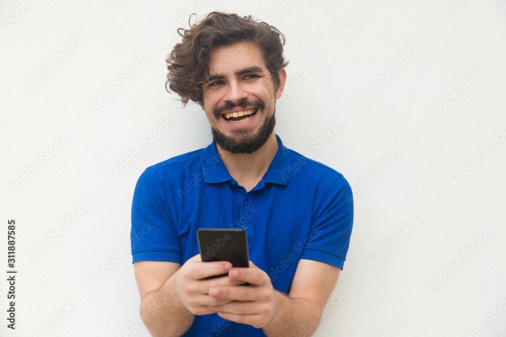 Joyful guy with cellphone getting funny news, laughing, looking at camera. Handsome bearded young man in blue casual t-shirt posing isolated over white background. Smartphone using concept Joyful guy with cellphone getting funny news, laughing, looking at camera. Handsome bearded young man in blue casual t-shirt posing isolated over white background. Smartphone using concept