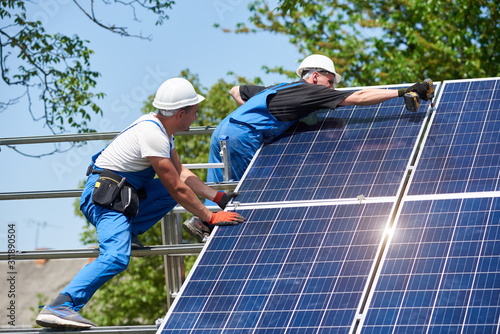 Two young technicians mounting with screwdriver heavy solar photo voltaic panel on tall steel platform on green tree background. Exterior solar panel voltaic system installation, dangerous job concept