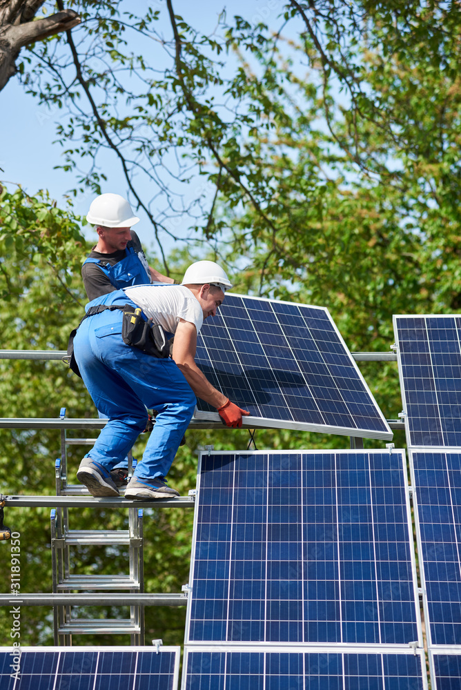 Two workers mounting heavy solar photo voltaic panel on tall steel platform standing on ladder on green tree and blue sky background. Exterior solar system installation, dangerous job concept. Two workers mounting heavy solar photo voltaic panel on tall steel platform standing on ladder on green tree and blue sky background. Exterior solar system installation, dangerous job concept.