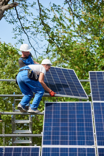 Two workers mounting heavy solar photo voltaic panel on tall steel platform standing on ladder on green tree and blue sky background. Exterior solar system installation, dangerous job concept.
