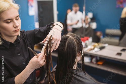 hairdresser holding scissors and comb and makes haircut woman client. Young beautiful woman hair cut in beauty salon. Process of hair cutting with use scissors. Hairdresser trimming brown.
