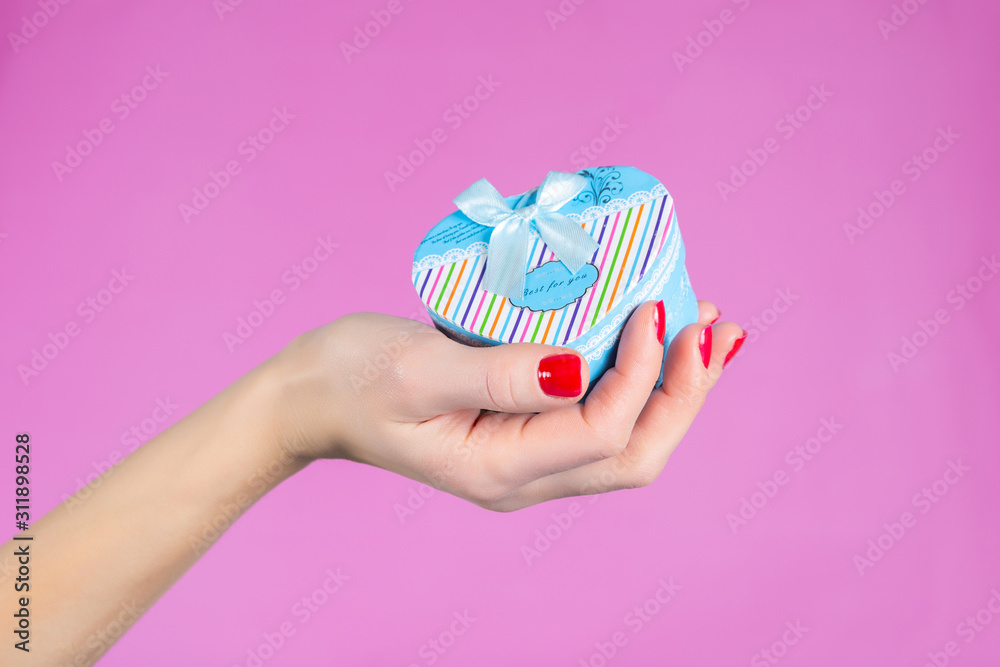 Female hand holding a blue gift box in heart shape isolated on pink background in studio. Female hands with red nails polish on fingers. Christmas or Valentines day holiday and minimalism concept Female hand holding a blue gift box in heart shape isolated on pink background in studio. Female hands with red nails polish on fingers. Christmas or Valentines day holiday and minimalism concept