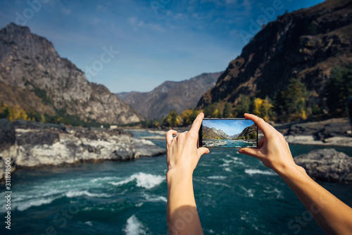 Hands hold a smartphone and take photo of mountain river and a rocks, close-up. Photographing the beautiful landscape on smartphone while traveling.