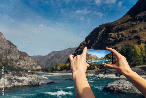 Hands hold a smartphone and take photo of mountain river and a rocks, close-up. Photographing the beautiful landscape on smartphone while traveling.