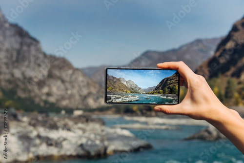 Female hand with smartphone close-up. Taking photo on smartphone while traveling. Delightful mountain landscape, stormy river, rocks and blue sky.