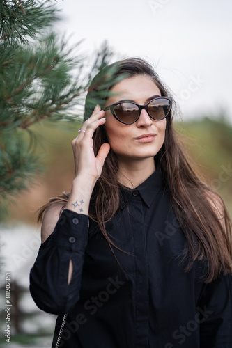 Obraz na płótnie Beautiful woman with brunette hair in dark clothes and sunglasses. Fashion street photography. Fashion model is posing on nature background. sunglasses shop w salonie