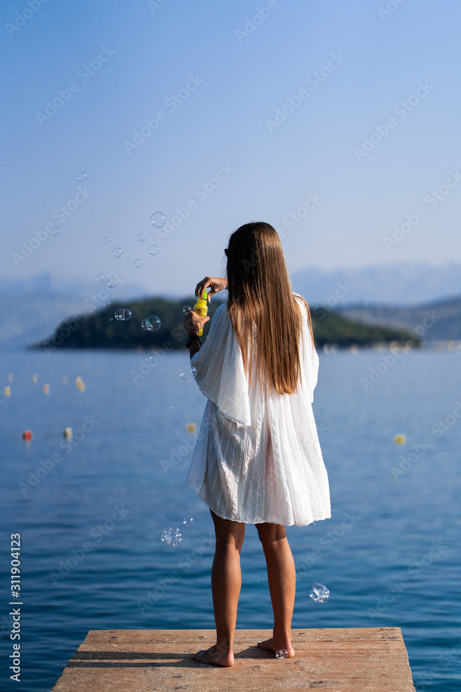 Young beautiful woman in white dress and sunglasses blowing soap bubbles pier with seaview background.. The concept of joy, and freedom during the vacation. The girl is enjoying the rest. Back view Young beautiful woman in white dress and sunglasses blowing soap bubbles pier with seaview background.. The concept of joy, and freedom during the vacation. The girl is enjoying the rest. Back view
