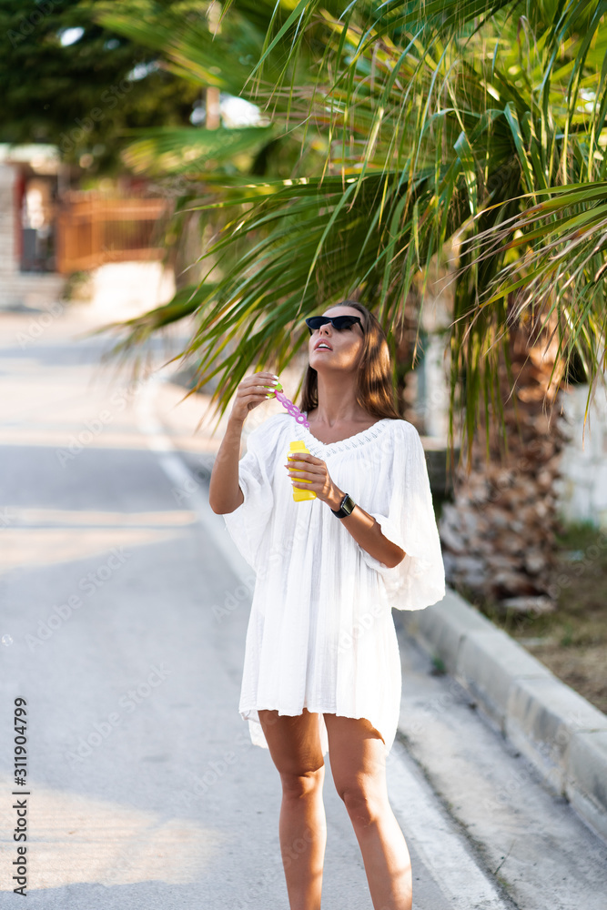 Young beautiful woman in white dress and sunglasses blowing soap bubbles on the road with palms. The concept of joy, ease and freedom during the vacation. The girl is enjoying the rest.