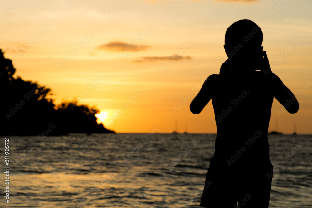 Silhouette portrait of young boy taking a seascape scene on the beach with light of sunset and blurred yacht boats on skyline in background