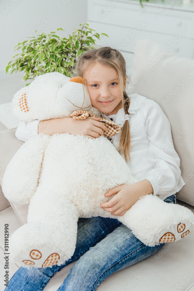 Happy little girl sitting on the sofa, hugging a big teddy-bear, smiling.