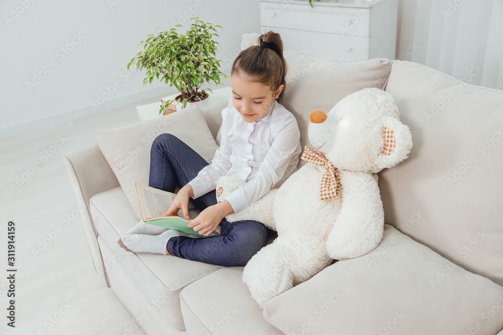 Cute little girl sitting on the sofa with teddy-bear near her, reading ...