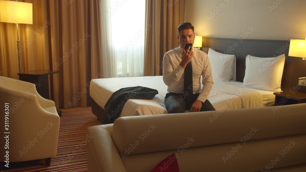Man in shirt and tie sit on the bed in hotel and record voice message on phone Man in shirt and tie sit on the bed in hotel and record voice message on phone