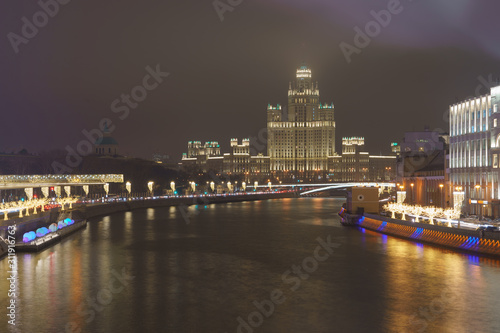 Photography of Stalinist skyscraper on Kotelnicheskaya embankment, big traffic, stret filled out cars and bridge in winter night. The spire hiding in the fog. International touristic concepts.