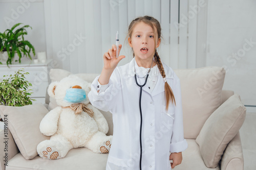 Fototapeta Naklejka Na Ścianę i Meble -  Smart little girl wearing doctor coat, stethoscope over neck, standing, holding a syringe with transparent liquid, looking at the camera.