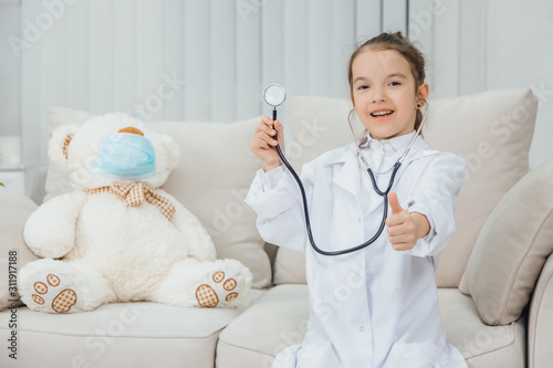 Little girl in medical coat holding a stethoscope, pointing her finger up. White teddy-bear in medical mask is sitting near her. photo