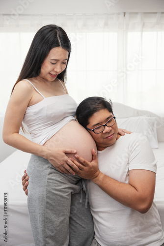 Handsome asia man is listening to his beautiful pregnant wife's tummy and smiling